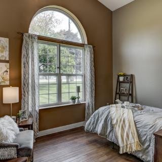 A cozy bedroom with a large window allowing natural light to fill the space, featuring wood flooring and stylish decor.