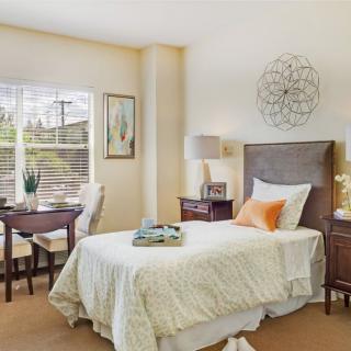 A cozy bedroom interior with a single bed, bedside tables, and a small dining area by the window.