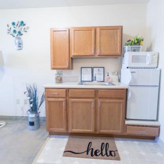 A compact kitchen setup with wooden cabinets, a small fridge, decorative plants, and a welcoming doormat.