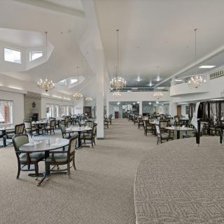 A spacious dining hall with multiple tables and chairs, decorated with chandeliers and carpeted flooring.