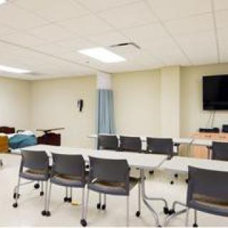 A hybrid classroom and medical training room with desks, chairs, a hospital bed, and a television.