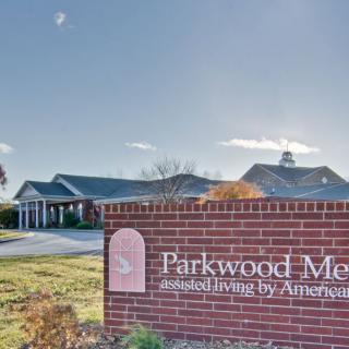 The image shows the entrance of Parkwood Meadows Residential Living, featuring a brick sign and part of the building in the background under a clear sky.