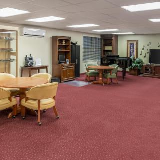 A spacious living room with red carpet, featuring various furniture including tables, chairs, a television, a piano, and shelves.