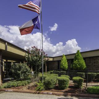 A view of a building's entrance with American and Texas flags flying, surrounded by well-maintained landscaping under a clear blue sky.