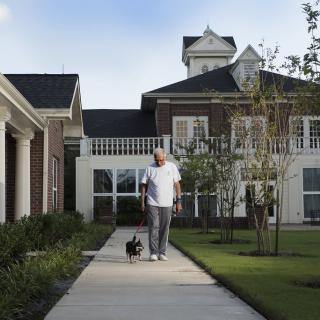 A man walks his dog along a sidewalk in front of a large house.