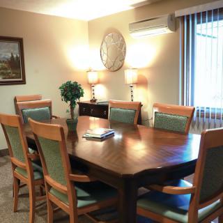 A well-lit dining room with a large wooden table and several matching chairs, decorated with lamps and a potted plant.