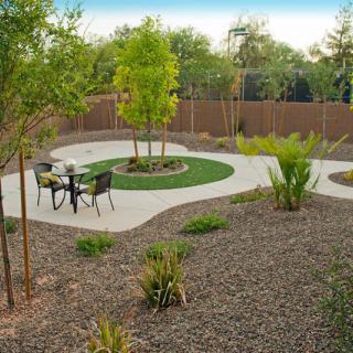 A landscaped garden with a patio area featuring outdoor seating, surrounded by trees, plants, and gravel paths.