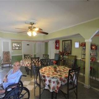 An elderly person in a wheelchair sits in a dining room with tables covered in floral tablecloths.