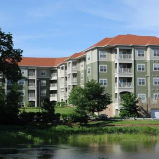 A residential apartment building with a green facade and red roof, situated near a pond and surrounded by trees.