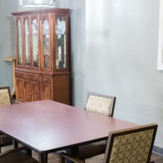 A dining room with a wooden table and chairs, and a display cabinet in the background.