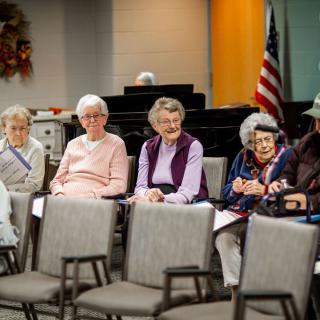 A group of elderly individuals seated in a meeting room with chairs arranged in rows.