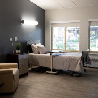 A modern and minimalist hospital room featuring a single bed with an adjacent chair, illuminated by natural light from large windows.