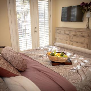 A cozy bedroom featuring a neatly made bed, a dresser with a television mounted above it, and natural light streaming through the shutters. A tray with fresh fruit adds a touch of hospitality to the space.