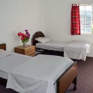 A simple bedroom with two twin beds, a wooden nightstand with flowers, and a window with red curtains.