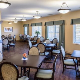 A well-lit dining area with multiple tables and chairs arranged neatly, featuring large windows with curtains.