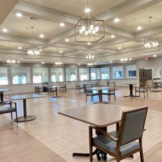 A spacious dining hall with multiple tables and chairs, illuminated by chandeliers and natural light from large windows.