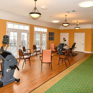 A well-lit exercise room with fitness equipment and chairs, featuring wooden floors and large windows.
