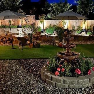 A beautifully lit garden at night featuring a central fountain, surrounded by flowers and outdoor seating areas.