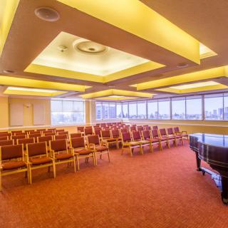 A spacious conference room with rows of seating, featuring a grand piano and large windows showcasing a cityscape.