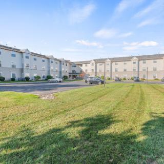 A view of a modern apartment building with a well-maintained lawn and parking area under a clear blue sky.
