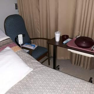 A view of a hospital patient room featuring a bed, chair, and bedside table.