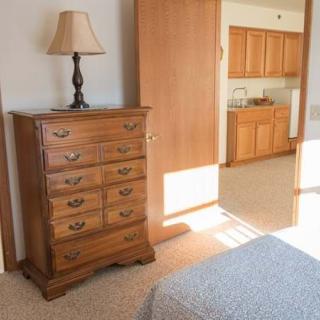 A cozy bedroom featuring a wooden dresser with a lamp on top, next to an open doorway leading to a kitchen area.
