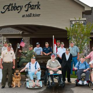 A group of veterans and supporters gathered outside Abbey Park at Mill River, displaying American flags and wearing military uniforms.