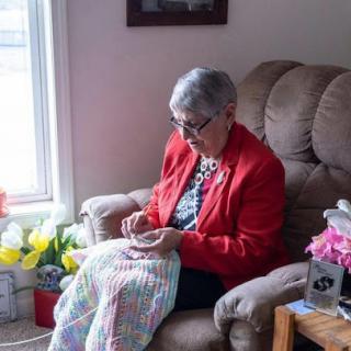 An elderly woman enjoys a cozy afternoon knitting in her comfortable chair, surrounded by vibrant flowers.