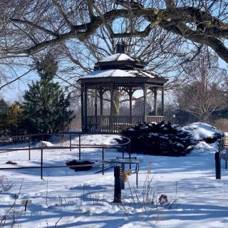 A picturesque gazebo stands amid a snowy winter landscape, surrounded by trees and enclosed by a fence.