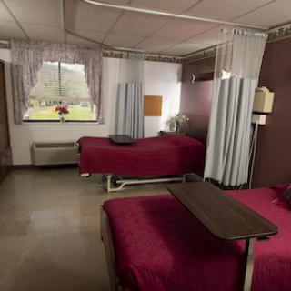 A hospital room with two beds separated by curtains, featuring maroon bedspreads and a window view.