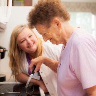 A caregiver assists an elderly woman with cooking in the kitchen.
