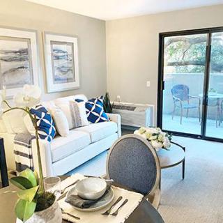 A modern living room featuring a white sofa with blue cushions, a dining table set, and large sliding glass doors opening to an outdoor area.