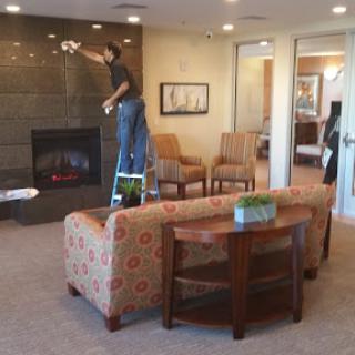 A man is painting a wall in a cozy living room with a fireplace, surrounded by comfortable seating.