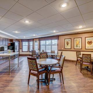 A spacious dining area with multiple tables and chairs, featuring a kitchen area in the background and decorative paintings on the walls.