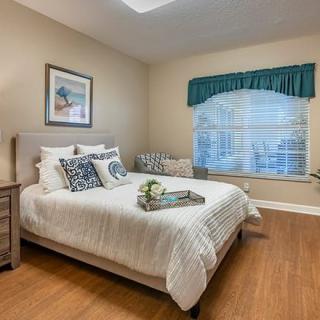 A cozy bedroom with a neatly made bed, wooden flooring, and a large window with green curtains.