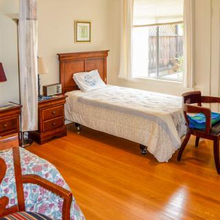 A cozy bedroom with a single bed and wooden furniture, featuring a nightstand, lamp, and a window letting in natural light.