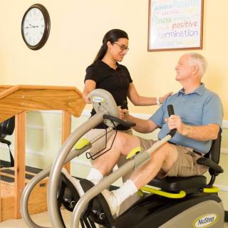 An elderly man receives guidance from a trainer while using an exercise machine for rehabilitation.