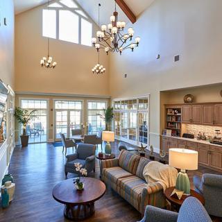 A spacious living room with high ceilings, elegant chandeliers, and large windows allowing natural light to flood the space. The room features comfortable seating and a warm, inviting decor.