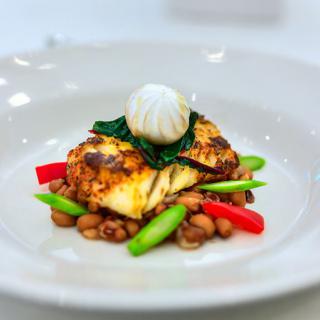 A gourmet dish featuring a grilled protein served with vibrant vegetables and beans, topped with a decorative garnish.