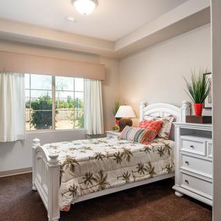 A well-lit bedroom with a neatly made bed, bedside table, and a large window.