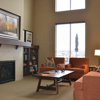 A cozy living room with a fireplace, orange armchairs, and a sofa centered around a wooden coffee table. Large windows provide natural light, enhancing the warm atmosphere.