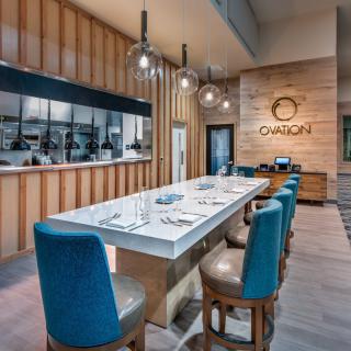 A modern restaurant interior featuring a long dining table with blue chairs and an open kitchen.