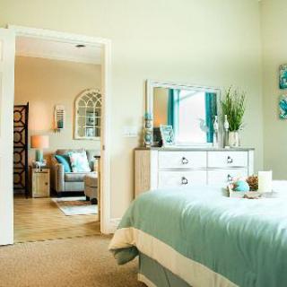 A cozy bedroom with a light and airy interior design, featuring a bed with teal bedding, a dresser with a mirror, and decorative plants.
