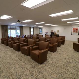 A spacious conference room with brown chairs arranged in rows, well-lit by overhead lights, and featuring large windows on one side.