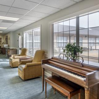 A cozy lounge area featuring a piano and comfortable chairs with ample natural light from large windows.