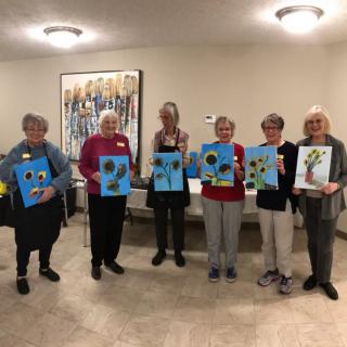 A group of seniors proudly displaying their flower paintings in an indoor setting.