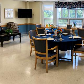 A dining room with a round table set for a meal, featuring several chairs, a wall-mounted TV, and large windows letting in natural light.