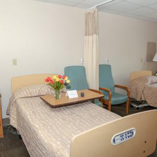 A hospital room with a bed, bedside table with flowers, and chairs.
