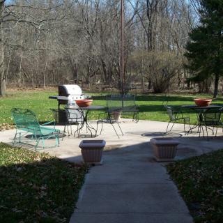 A serene outdoor patio area with garden furniture and a grill, surrounded by lush greenery.