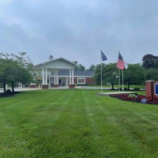 A building surrounded by lush greenery with flags and a sign in the foreground on a cloudy day.